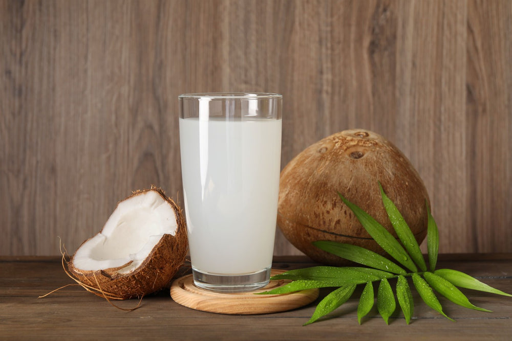 A glass of coconut water sits next to a sliced open coconut and palm leaf.