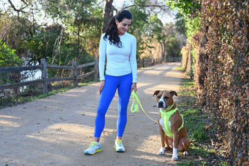 A woman dressed in athletic gear stands on a running trail with her dog.