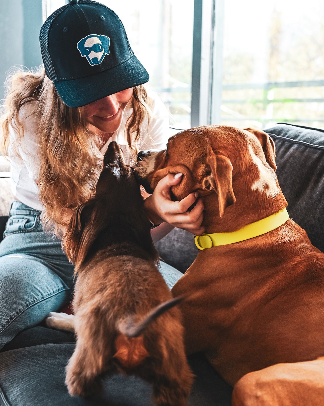 A woman smiling and holding her face up to a pit bull. There is a small weiner dog on the couch to the left of the pit bull.She is wearing a Native Pet hat