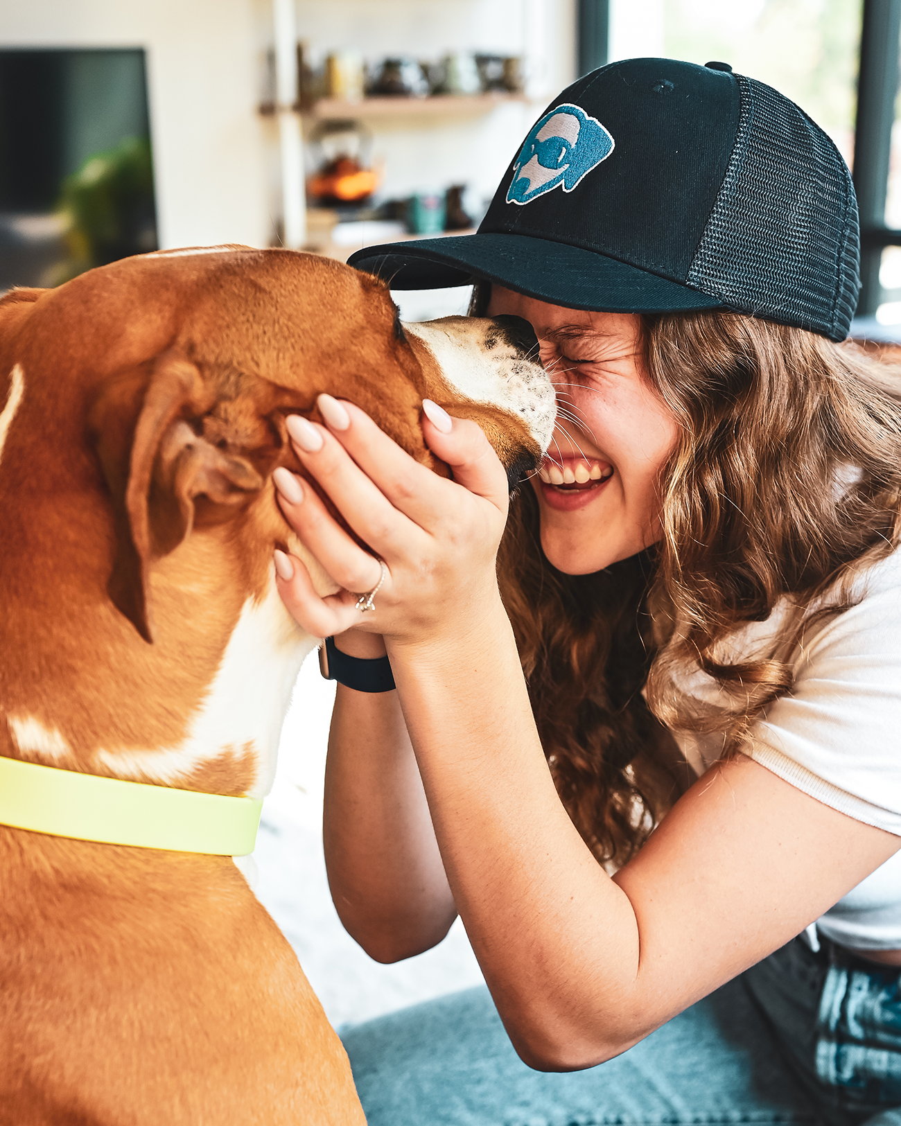 A woman smiling and holding her face up to a pit bull. She is wearing a Native Pet hat